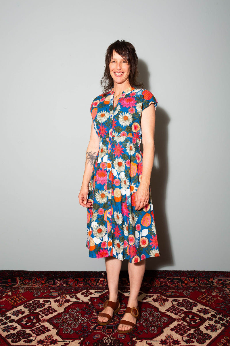 Woman wearing a colorful floral dress standing on a patterned rug with a plain wall background, Woman wearing a patterned dress standing on a rug,  Sapling Textiles, Fabric Store Australia, Indian Fabric online Australia