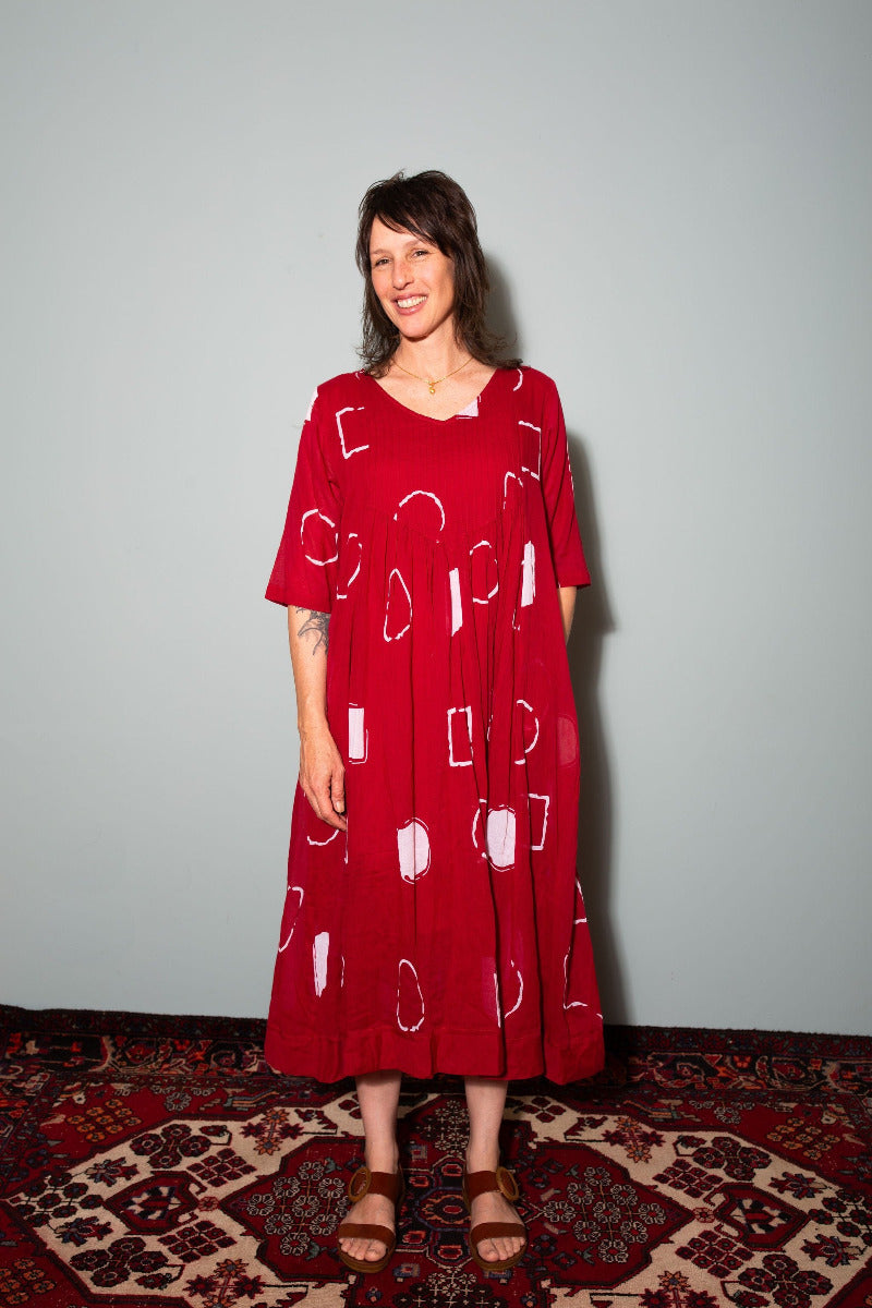 Woman wearing a red dress with white patterns on a plain background. Woman wearing a patterned dress standing on a rug against a plain background,  Sapling Textiles, Fabric Store Australia, Indian Fabric online Australia