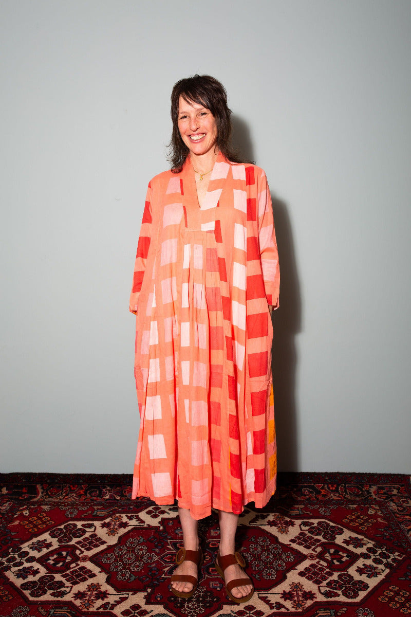Woman wearing a colorful patterned dress standing on a patterned rug with a plain wall background. Woman wearing a patterned dress standing on a rug against a plain background,  Sapling Textiles, Fabric Store Australia, Indian Fabric online Australia