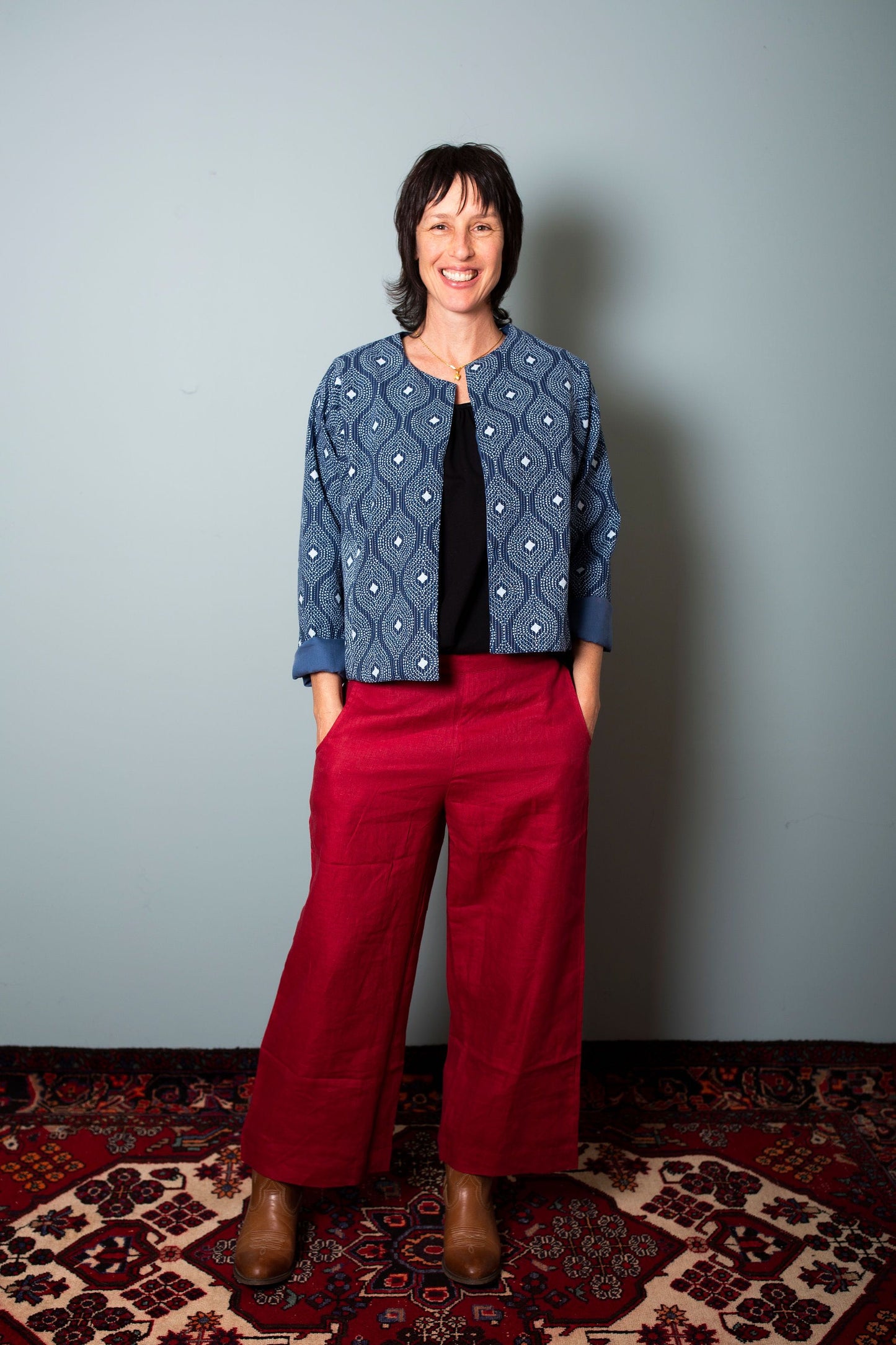 A person standing against a grey background, wearing a red garment with a geometric pattern on the top and a black shirt underneath. They are also wearing a blue and indigo jacket with a round neckline. Indian fabric, block print fabric, Indian block print fabric, Indian woodblock print fabric, Indian cotton fabric